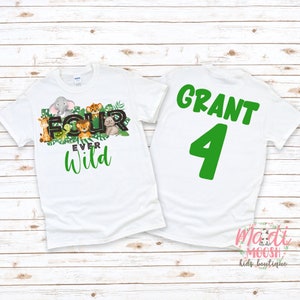 May include: White t-shirts with a jungle theme. One shirt features the text "FOUR EVER WILD" with animal illustrations. The other shirt has the name "GRANT" and the number "4" in green. The shirts are on a light wood background.