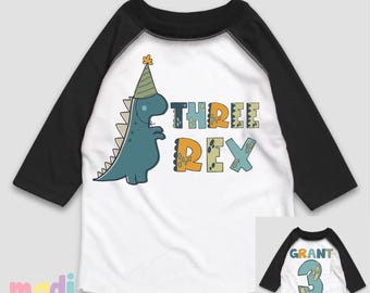 May include: White and black raglan-style toddler shirt with a blue dinosaur graphic wearing a party hat. The shirt says "THREE REX" in colorful letters. The back of the shirt has the name "GRANT" and the number "3".