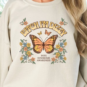 May include: A cream colored sweatshirt with the text "Bitch I'm Dizzy" in a yellow and orange font with a butterfly graphic and the text "POTS Syndrome Awareness" below the butterfly.