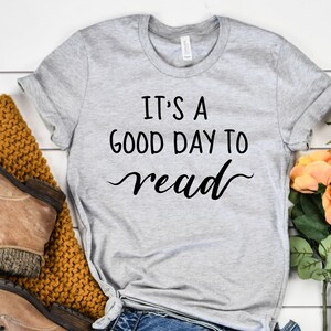 May include: A gray t-shirt with the text "It's a good day to read" in black script. The shirt is laid out on a white wooden surface with a pair of brown boots, a knitted sweater, and a bouquet of peach and white roses.