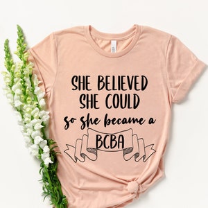 Pode incluir: Uma camiseta cor de pêssego com o texto "She believed she could so she became a BCBA" impresso em preto.