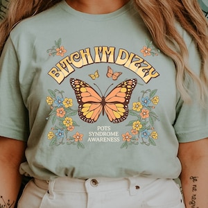 Puede incluir: Camiseta verde claro con el texto "Bitch I'm Dizzy" sobre una mariposa. El diseño incluye flores y las palabras "POTS Syndrome Awareness". La camiseta es de estilo casual.
