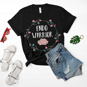 May include: Black t-shirt with a floral wreath design and the text "ENDO WARRIOR" in white. The shirt is paired with denim shorts and sandals.