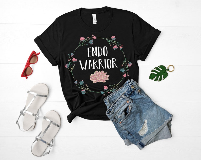 May include: Black t-shirt with a floral wreath design and the text "ENDO WARRIOR" in white. The shirt is paired with denim shorts and sandals.