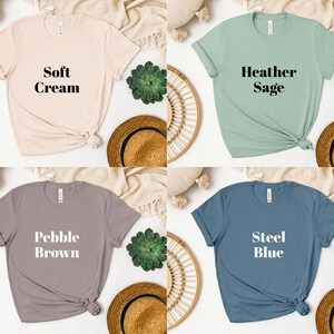 May include: Four t-shirts in different colors with text on the front. The colors are soft cream, heather sage, pebble brown, and steel blue. The text on the shirts reads "Soft Cream", "Heather Sage", "Pebble Brown", and "Steel Blue".