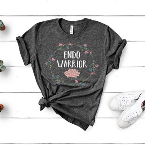 May include: A dark gray t-shirt with a floral wreath design and the text "Endo Warrior" in white.