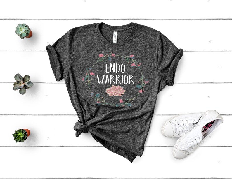 May include: A dark gray t-shirt with a floral wreath design and the text "Endo Warrior" in white.