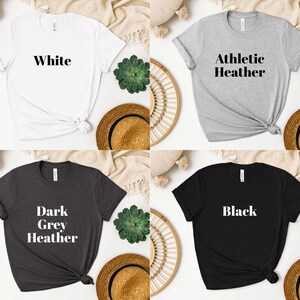 May include: Four t-shirts in different colors: white, athletic heather, dark grey heather, and black. Each shirt has a different text printed on the front: "White", "Athletic Heather", "Dark Grey Heather", and "Black".