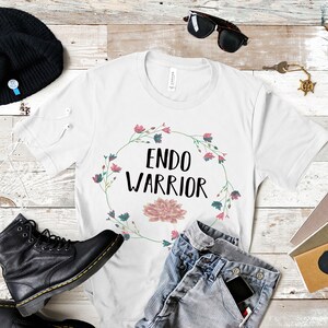 May include: White t-shirt with a floral wreath design and the text "ENDO WARRIOR" in black lettering. The shirt is laid flat on a wooden surface with a pair of black boots, a beanie, sunglasses, and a pair of jeans.