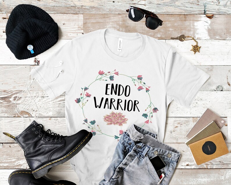 May include: White t-shirt with a floral wreath design and the text "ENDO WARRIOR" in black lettering. The shirt is laid flat on a wooden surface with a pair of black boots, a beanie, sunglasses, and a pair of jeans.