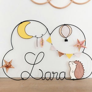 May include: A black wire frame in the shape of a cloud with the name "Lara" written in cursive. The frame is decorated with a yellow crescent moon, a hot air balloon, a hedgehog, and origami stars.