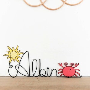 May include: A black wire name art piece with the name "Albin" spelled out. A yellow sun and a red crab are attached to the wire.
