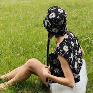 Pu&ograve; includere: Un cappello a secchiello nero con ricamo floreale bianco e rosso. Il cappello ha un nastro nero legato attorno alla corona. La persona che indossa il cappello &egrave; seduta su un prato erboso.