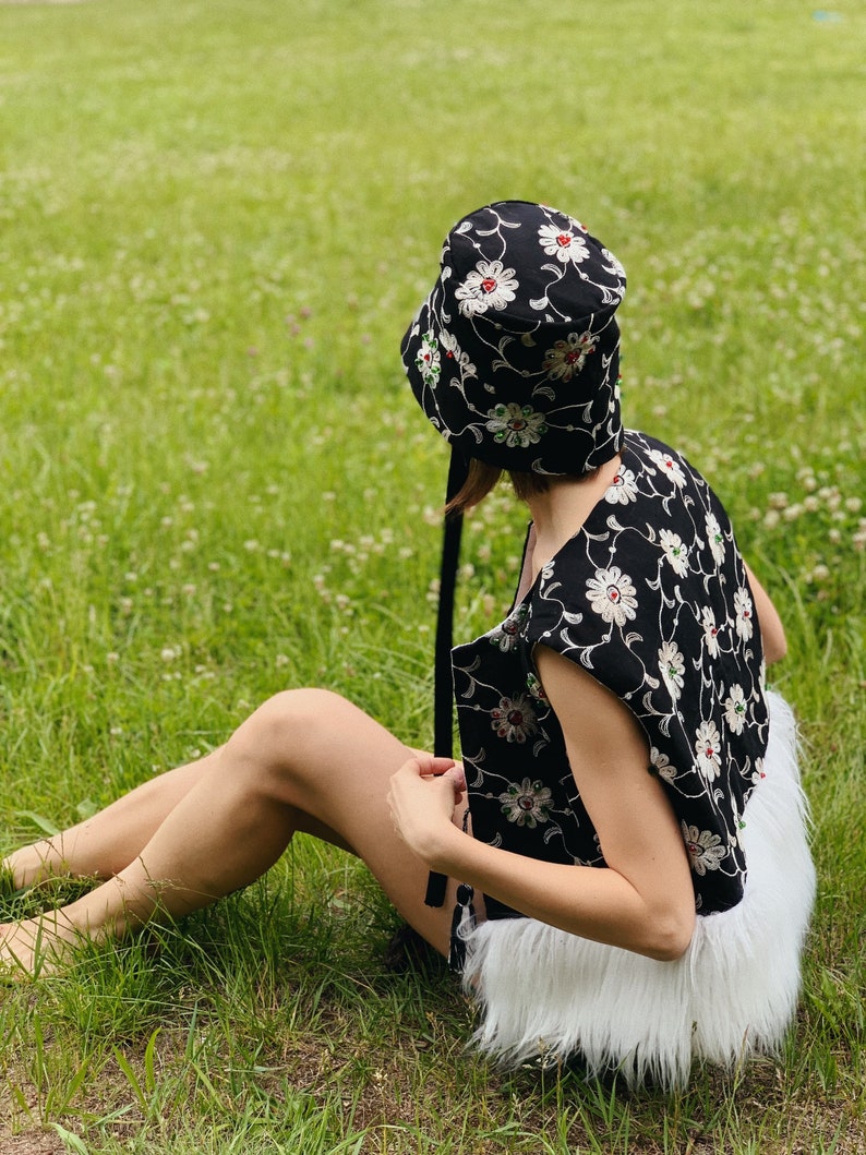 Pu&ograve; includere: Un cappello a secchiello nero con ricamo floreale bianco e rosso. Il cappello ha un nastro nero legato attorno alla corona. La persona che indossa il cappello &egrave; seduta su un prato erboso.