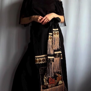 Peut inclure: Robe noire à épaules dénudées avec franges décoratives et un panneau brodé détaillé. La robe a une attache à la taille et des bretelles délicates. Le panneau brodé représente des personnages dans une scène aux tons chauds.