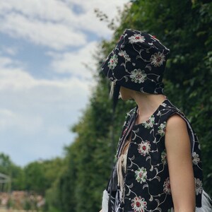 Pu&ograve; includere: Un gilet nero con ricami floreali bianchi e rossi. Il gilet &egrave; senza maniche e viene indossato sopra una camicia bianca con una sciarpa a motivi bianchi e neri. La persona indossa un cappello nero con ricami floreali bianchi e rossi.