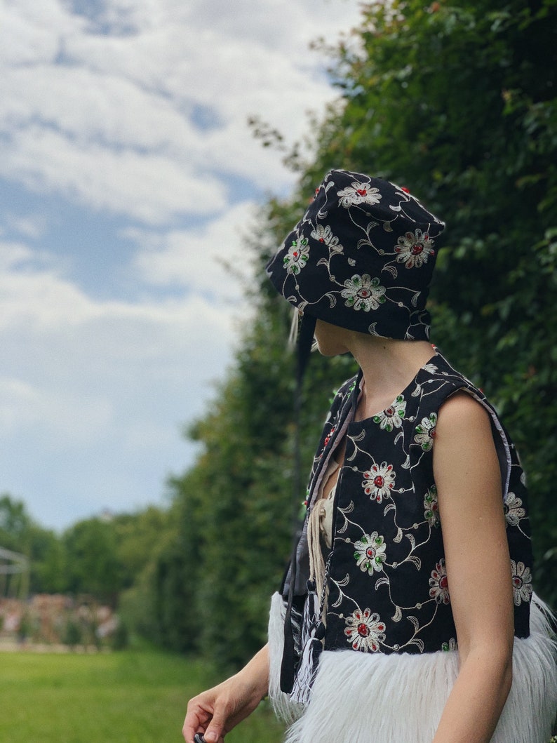 Pu&ograve; includere: Un gilet nero con ricami floreali bianchi e rossi. Il gilet &egrave; senza maniche e viene indossato sopra una camicia bianca con una sciarpa a motivi bianchi e neri. La persona indossa un cappello nero con ricami floreali bianchi e rossi.