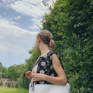 Pu&ograve; includere: Una donna indossa un gilet nero senza maniche con ricami floreali e un bordo in pelliccia sintetica bianca. Il gilet ha un dettaglio a frangia sul fondo. &Egrave; in piedi in un'area erbosa con una siepe verde alle sue spalle.