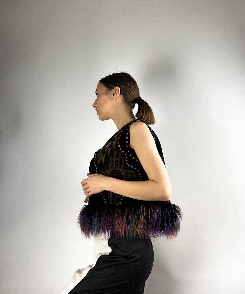 May include: A black vest with a geometric pattern and colorful fur trim. The vest has a dark background with gold and brown accents. The fur trim features shades of purple, blue, orange, and brown. The vest is paired with black and white pants.
