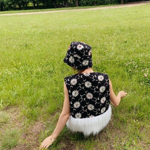 Pu&ograve; includere: Una persona indossa un gilet nero e bianco a fiori con un bordo in pelliccia bianca e un cappello a secchiello nero e bianco a fiori. La persona &egrave; seduta su un prato erboso.