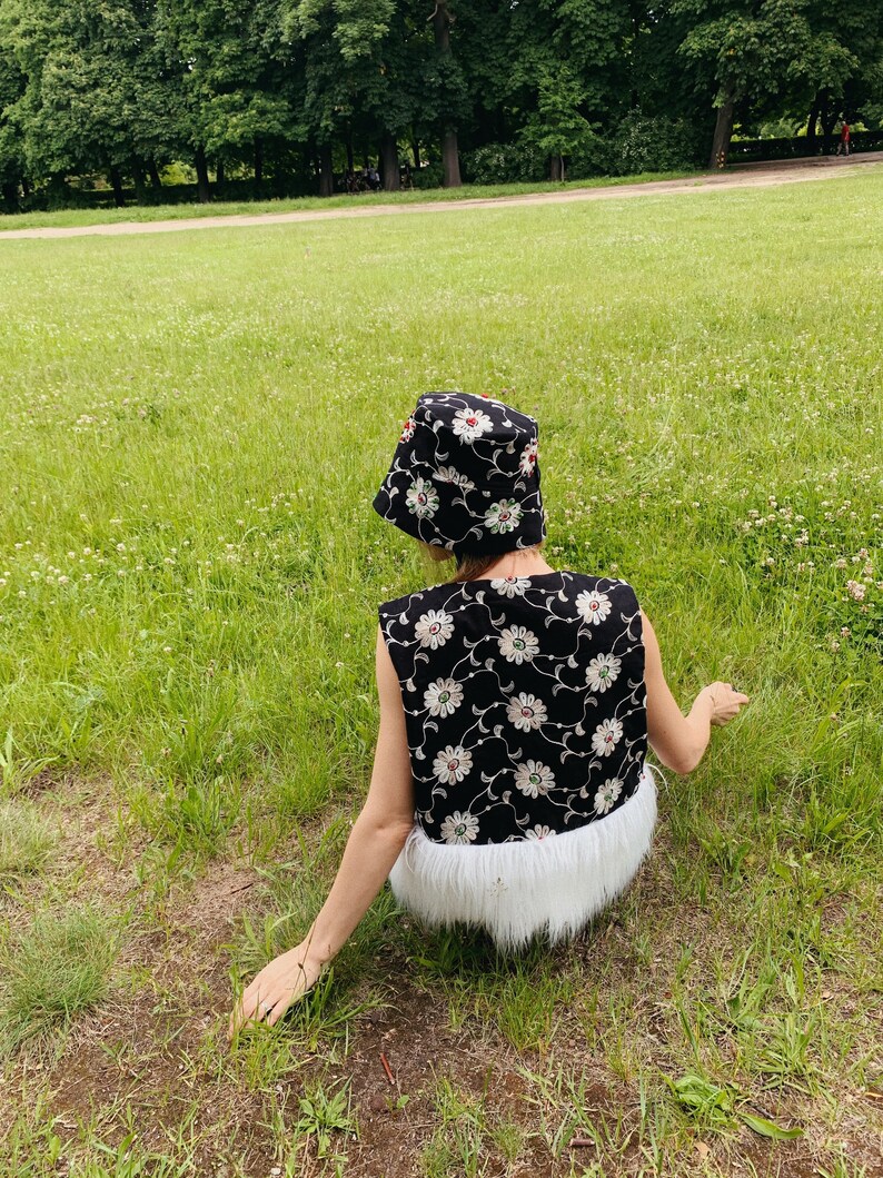 Pu&ograve; includere: Una persona indossa un gilet nero e bianco a fiori con un bordo in pelliccia bianca e un cappello a secchiello nero e bianco a fiori. La persona &egrave; seduta su un prato erboso.