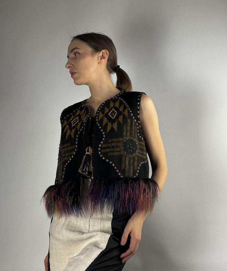 May include: A black vest with geometric patterns in brown and gold, embellished with small beads and a tassel. The vest features colorful faux fur trim in shades of purple, blue, and orange. The vest is sleeveless.