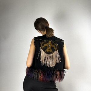 May include: Black vest with a decorative design on the back. The design features a central emblem with fringe and star-shaped accents. The bottom of the vest has a colorful fur trim.