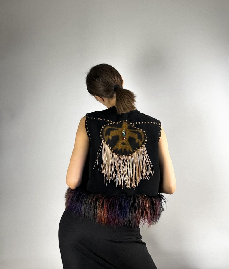 May include: Black vest with a decorative design on the back. The design features a central emblem with fringe and star-shaped accents. The bottom of the vest has a colorful fur trim.