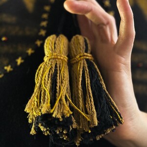 May include: Two decorative tassels with a mix of gold and black yarn. The tassels are held in a hand, with a dark background. The tassels are tied with a gold-colored thread and have a beaded detail.