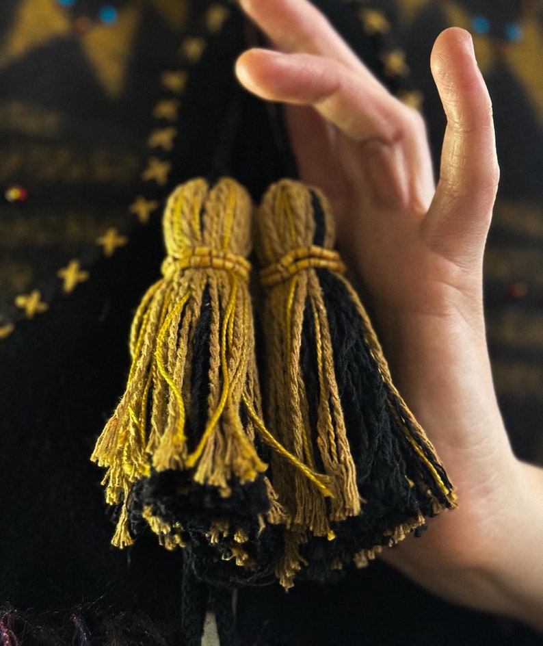 May include: Two decorative tassels with a mix of gold and black yarn. The tassels are held in a hand, with a dark background. The tassels are tied with a gold-colored thread and have a beaded detail.