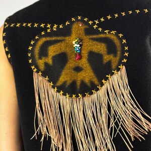 May include: Black vest with a gold-colored Native American-inspired design on the back. The design is outlined with gold stitching and features a beaded embellishment. Long, thin, gold-colored fringe hangs from the bottom.