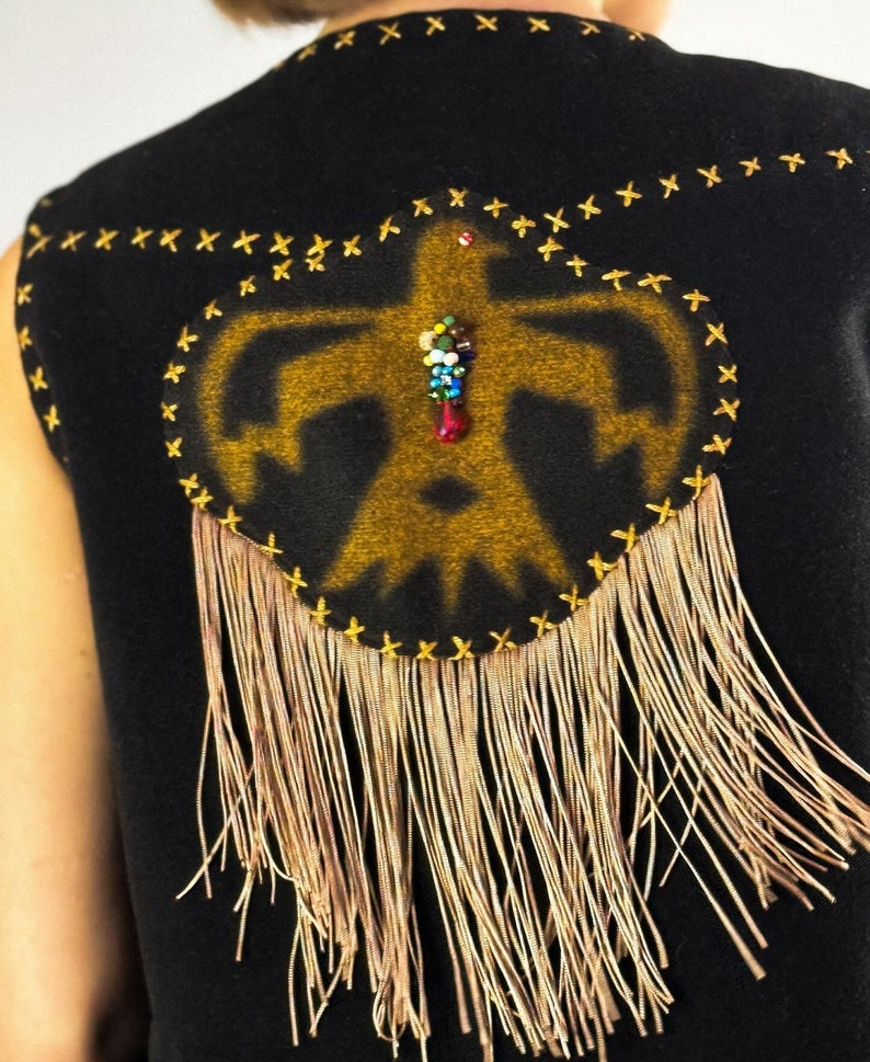 May include: Black vest with a gold-colored Native American-inspired design on the back. The design is outlined with gold stitching and features a beaded embellishment. Long, thin, gold-colored fringe hangs from the bottom.