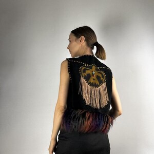 May include: Black sleeveless vest with a decorative design on the back. The design features a gold-colored emblem with fringe and colorful fur trim at the bottom. The vest is made of a dark material.