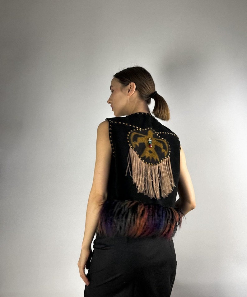 May include: Black sleeveless vest with a decorative design on the back. The design features a gold-colored emblem with fringe and colorful fur trim at the bottom. The vest is made of a dark material.