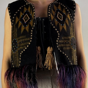 May include: A black vest with a geometric pattern in shades of brown and gold, embellished with small beads and decorative stitching. The vest features a fringe of colorful faux fur at the bottom and two brown tassels.