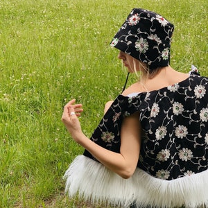 Pu&ograve; includere: Un cappello a secchiello nero con ricamo floreale bianco e rosso. Il cappello &egrave; indossato da una persona seduta su un campo erboso. Il cappello ha un bordo in pelliccia bianca.