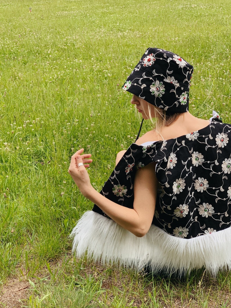 Pu&ograve; includere: Un cappello a secchiello nero con ricamo floreale bianco e rosso. Il cappello &egrave; indossato da una persona seduta su un campo erboso. Il cappello ha un bordo in pelliccia bianca.