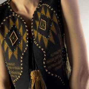 May include: Black vest with a geometric design in shades of gold and brown. The vest is adorned with small, colorful beads and decorative stitching. A tassel hangs from the side. The vest is sleeveless.