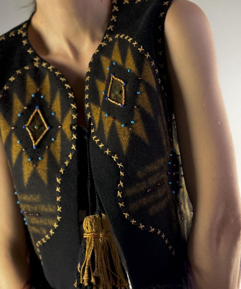 May include: Black vest with a geometric design in shades of gold and brown. The vest is adorned with small, colorful beads and decorative stitching. A tassel hangs from the side. The vest is sleeveless.