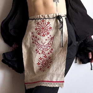 Peut inclure: Un tablier en lin beige avec des fleurs brodées rouges et un motif d'oiseau. Le tablier a un cordon de serrage noir et une bordure en dentelle blanche.