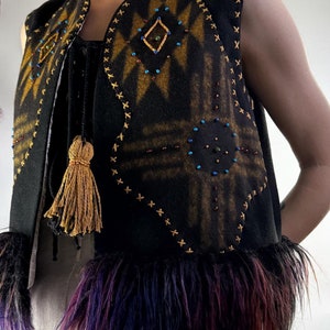 May include: Black vest with a geometric design in shades of gold and brown, embellished with colorful beads. The vest features a fringe of purple, red, and black fur at the bottom and a gold tassel. The edges are stitched with gold thread.
