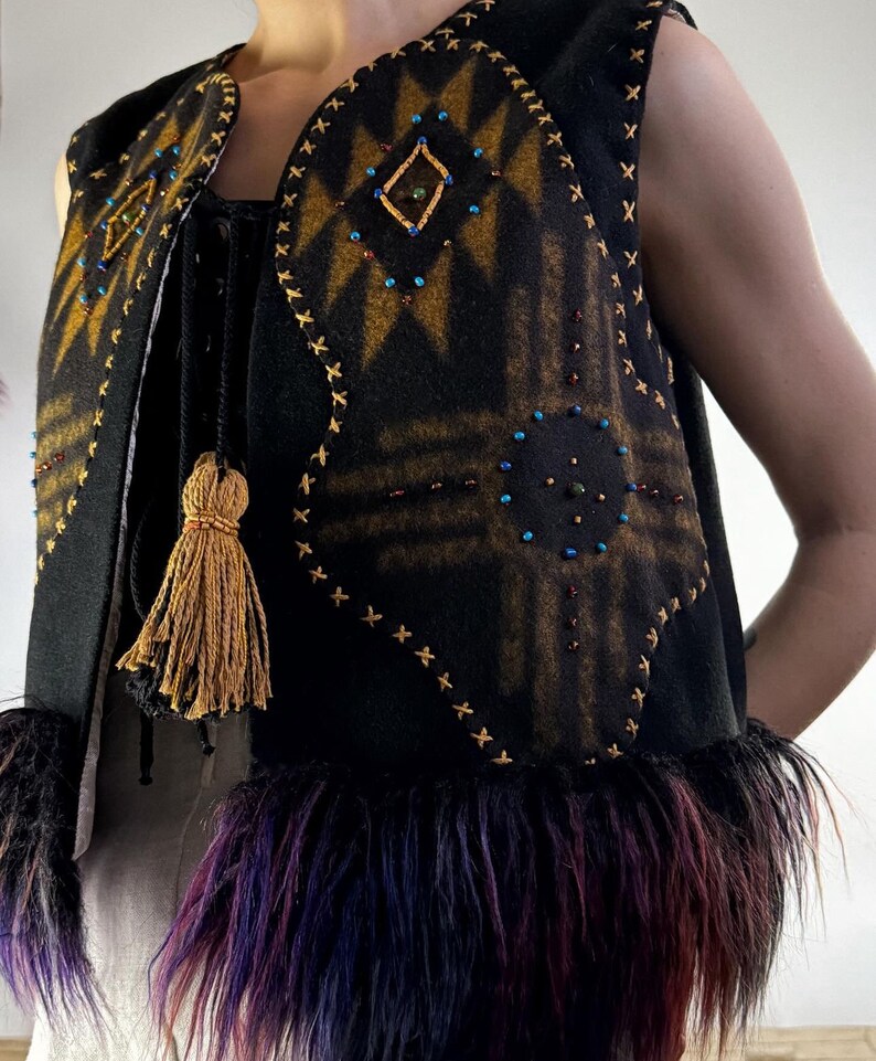 May include: Black vest with a geometric design in shades of gold and brown, embellished with colorful beads. The vest features a fringe of purple, red, and black fur at the bottom and a gold tassel. The edges are stitched with gold thread.