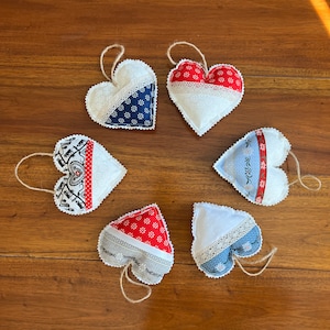 Puede incluir: Seis adornos hechos a mano en forma de corazón con diferentes patrones. Los adornos están hechos de tela blanca con patrones rojos, azules y blancos. Los adornos están colgados de una superficie de madera.