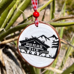 Puede incluir: Un adorno de madera blanco con una silueta negra de un chalet suizo con una cruz roja en el techo. El adorno está colgado de una cinta roja y blanca a cuadros.