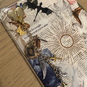 May include: A clear phone case with a decorative charm. The charm includes a gold heart, butterfly, and various silver and gold pendants, including a wing, cross, and dagger. The case has a collage design with text and floral elements.