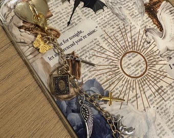 Könnte beinhalten: Eine transparente Handyhülle mit einem dekorativen Anhänger. Der Anhänger enthält ein goldenes Herz, einen Schmetterling und verschiedene silberne und goldene Anhänger, darunter ein Flügel, ein Kreuz und ein Dolch. Die Hülle hat ein Collage-Design mit Text und floralen Elementen.