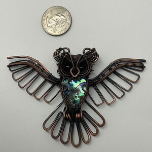 May include: A handcrafted copper wire owl pendant with outstretched wings. The owl features a central abalone shell and black bead eyes. A U.S. quarter coin is next to the owl for size comparison. The owl's wings are made of copper wire.