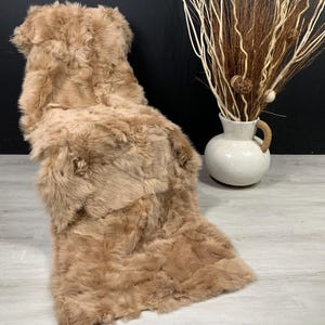 May include: A plush, tan-colored sheepskin rug. The rug is rectangular with a soft, textured surface. A white ceramic vase with a handle and decorative branches is in the background.
