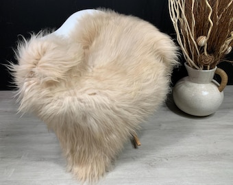 Könnte beinhalten: Ein hellbeige, flauschiger Schaffellteppich, der über einem weißen Stuhl drapiert ist. Der Teppich hat lange, weiche Fasern und eine natürliche, unregelmäßige Form. Rechts befindet sich eine dekorative Vase mit getrockneten Zweigen.