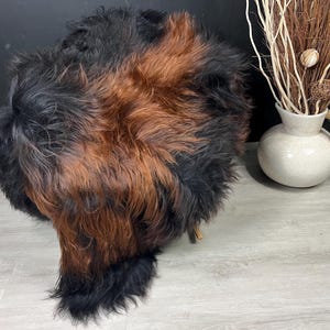 May include: A fluffy, long-haired sheepskin rug featuring a blend of deep black and rich reddish-brown fur. The texture is thick and shaggy, with the dark tones concentrated at the edges and the lighter brown hues integrated throughout the center.